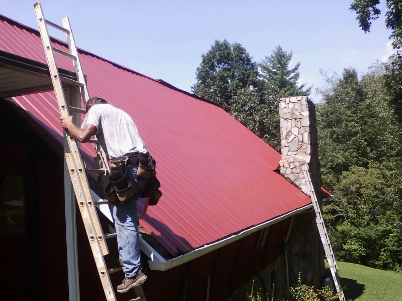 Roofer on a ladder beside a red metal roof and stone chimney — Soffit & Fascia Repair by Roofing Brothers in The Plains